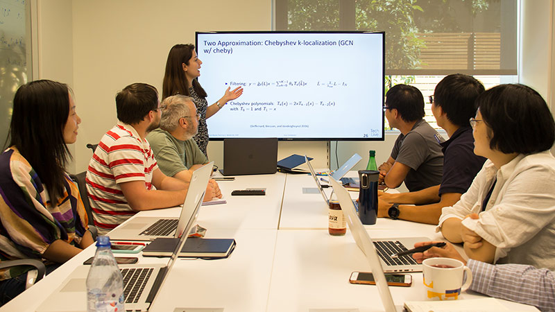 A group of people sitting around a conference table, watching a woman present something on screen.